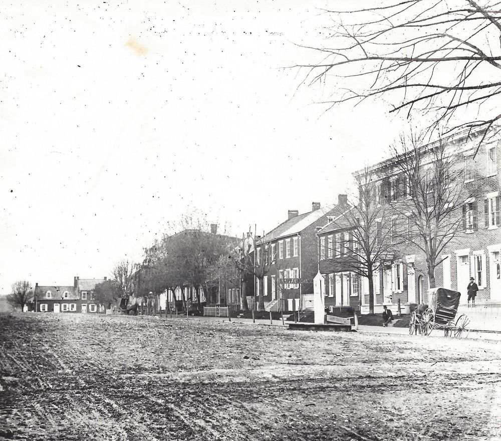 Black-and-white street scene with row houses, bare trees, and horse-drawn carriages along a muddy road