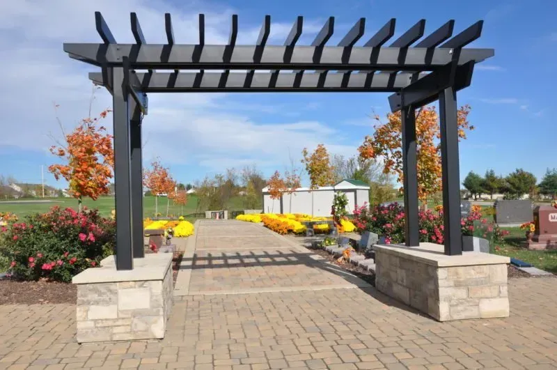 Black pergola entrance over a brick walkway in a landscaped garden with colorful flowers and autumn trees