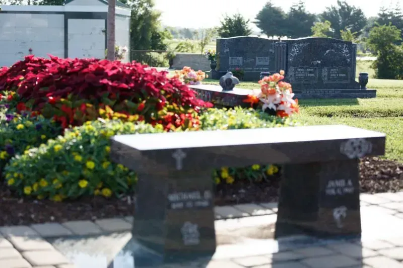 Memorial bench beside colorful flowers and gravestones in a sunny cemetery garden