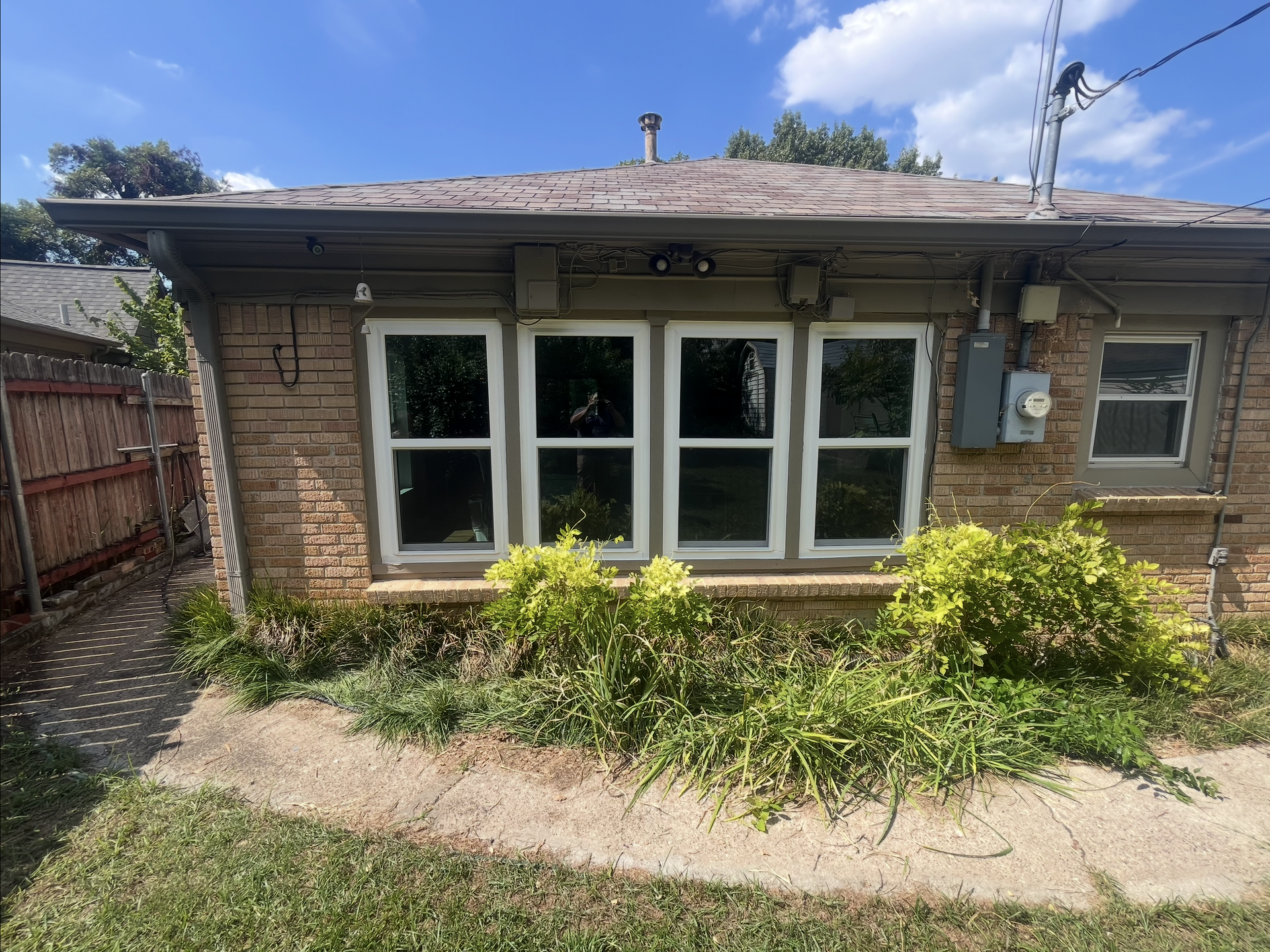 Brick building with white-framed windows, overgrown plants, and electrical box.