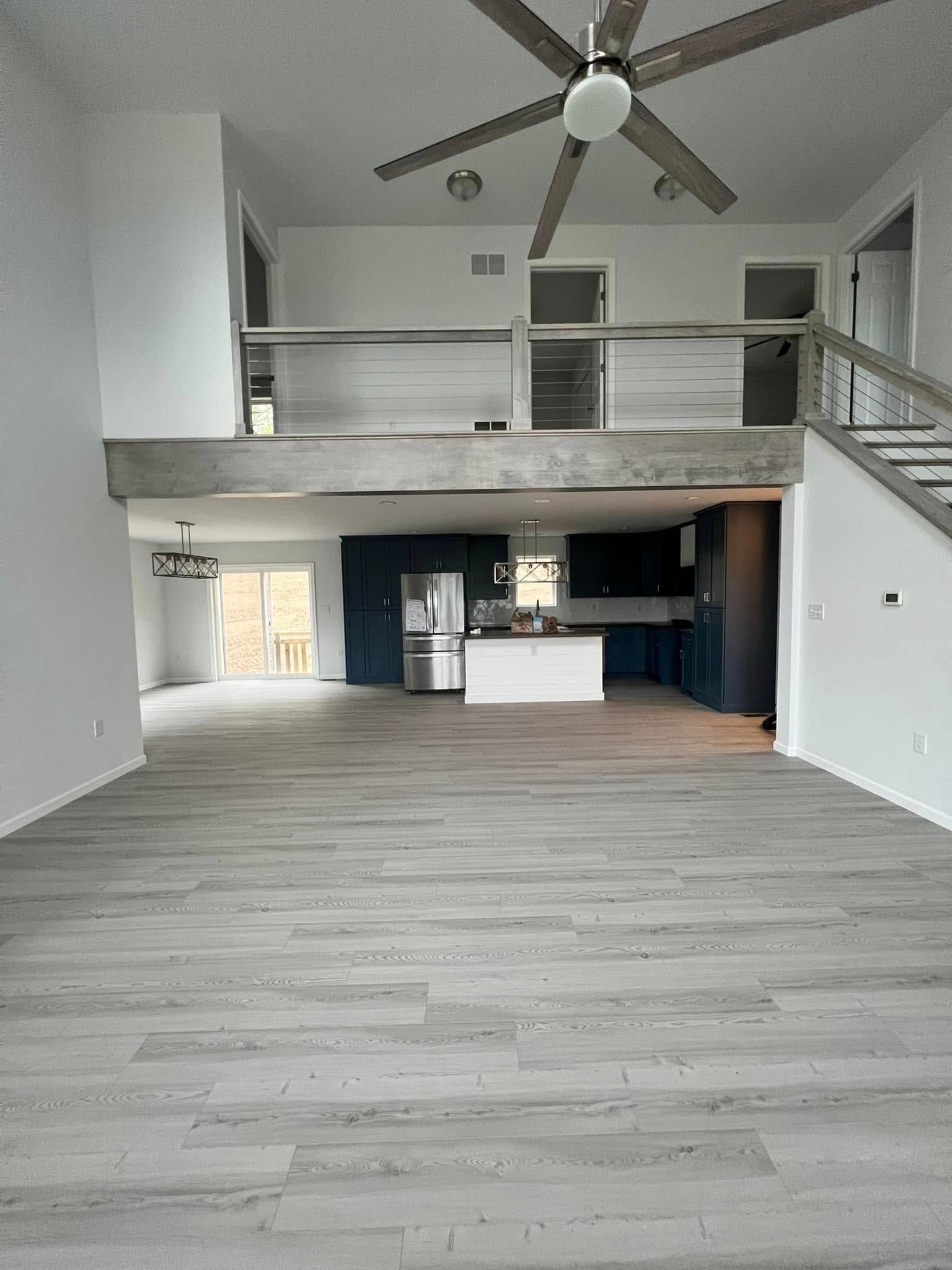 Empty modern loft with gray floors, high ceilings, ceiling fan, and kitchen area with staircase.