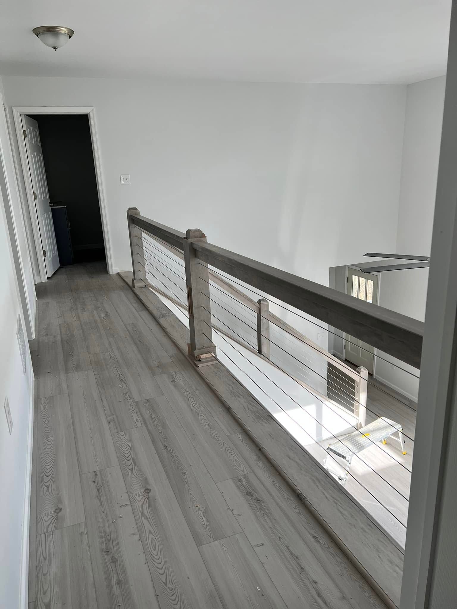Empty upstairs hallway with gray wood floor and white walls, beside a modern railing overlooking a lower level