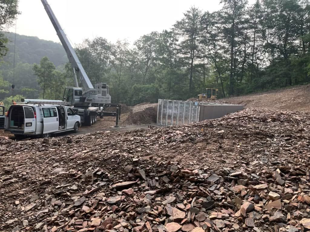 Crane and trucks at a construction site with rubble and retaining wall under trees