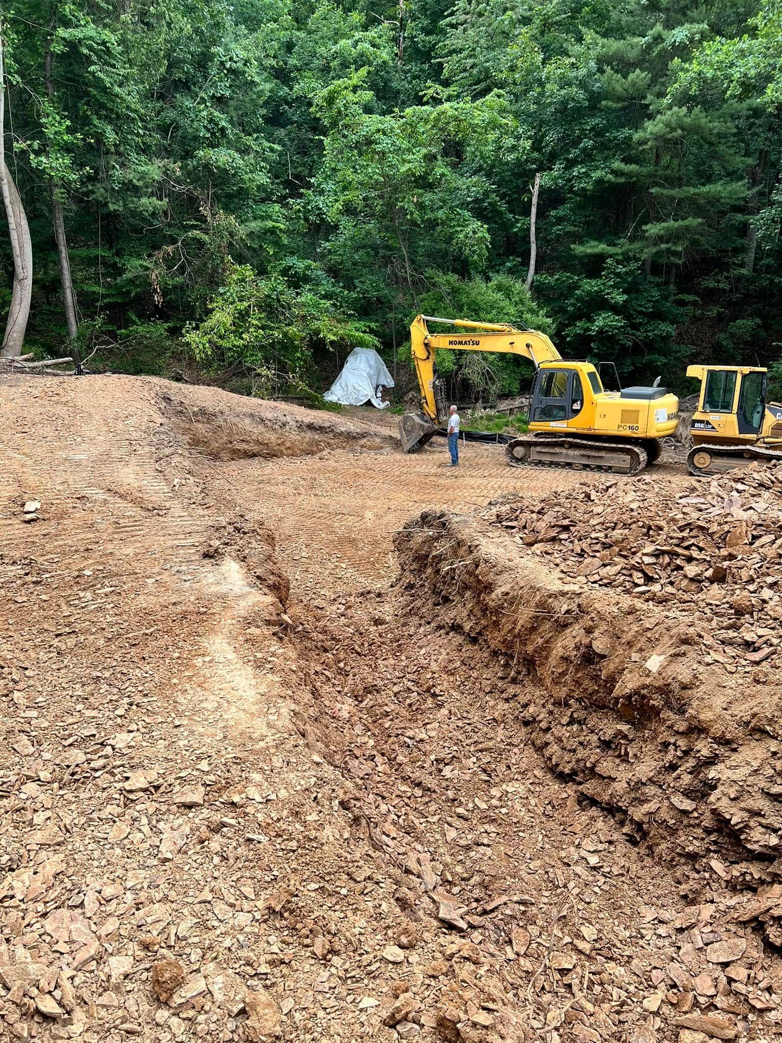 Cleared dirt path in a wooded area with two yellow excavators and a worker nearby