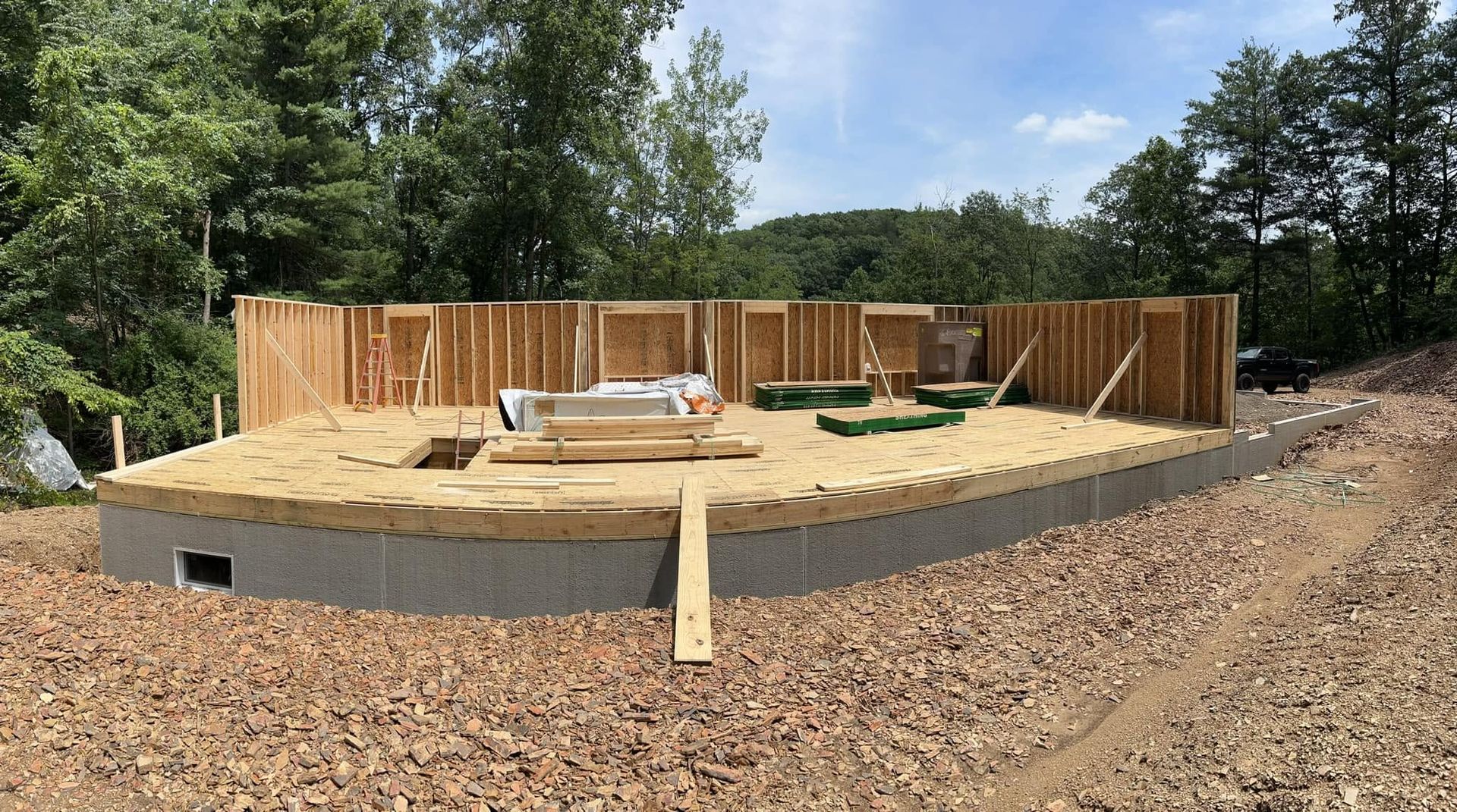 Partially built wooden house foundation with framing, lumber, and concrete wall in a wooded clearing