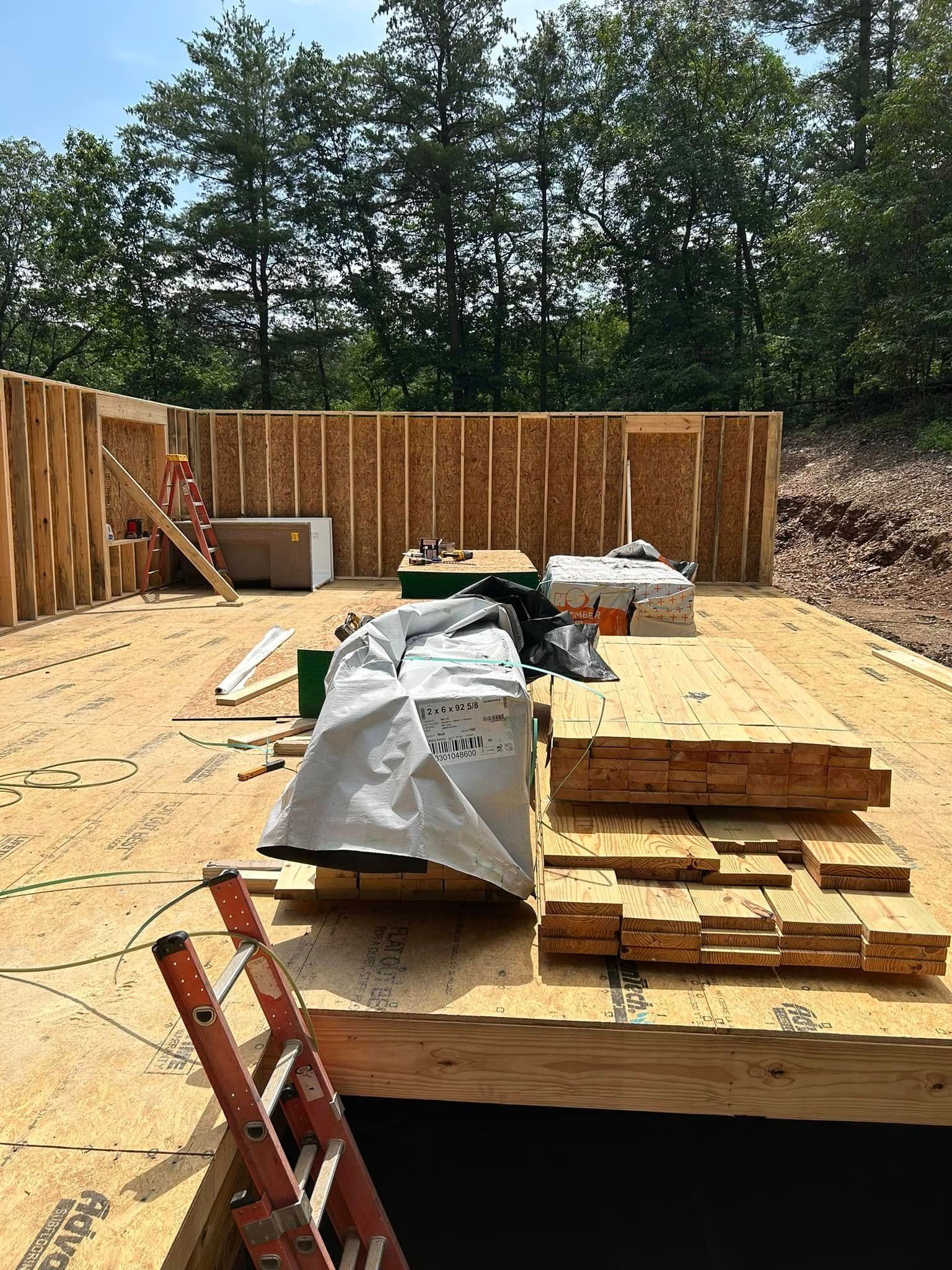 Partially built wooden house frame with plywood floor and construction materials under a tree line