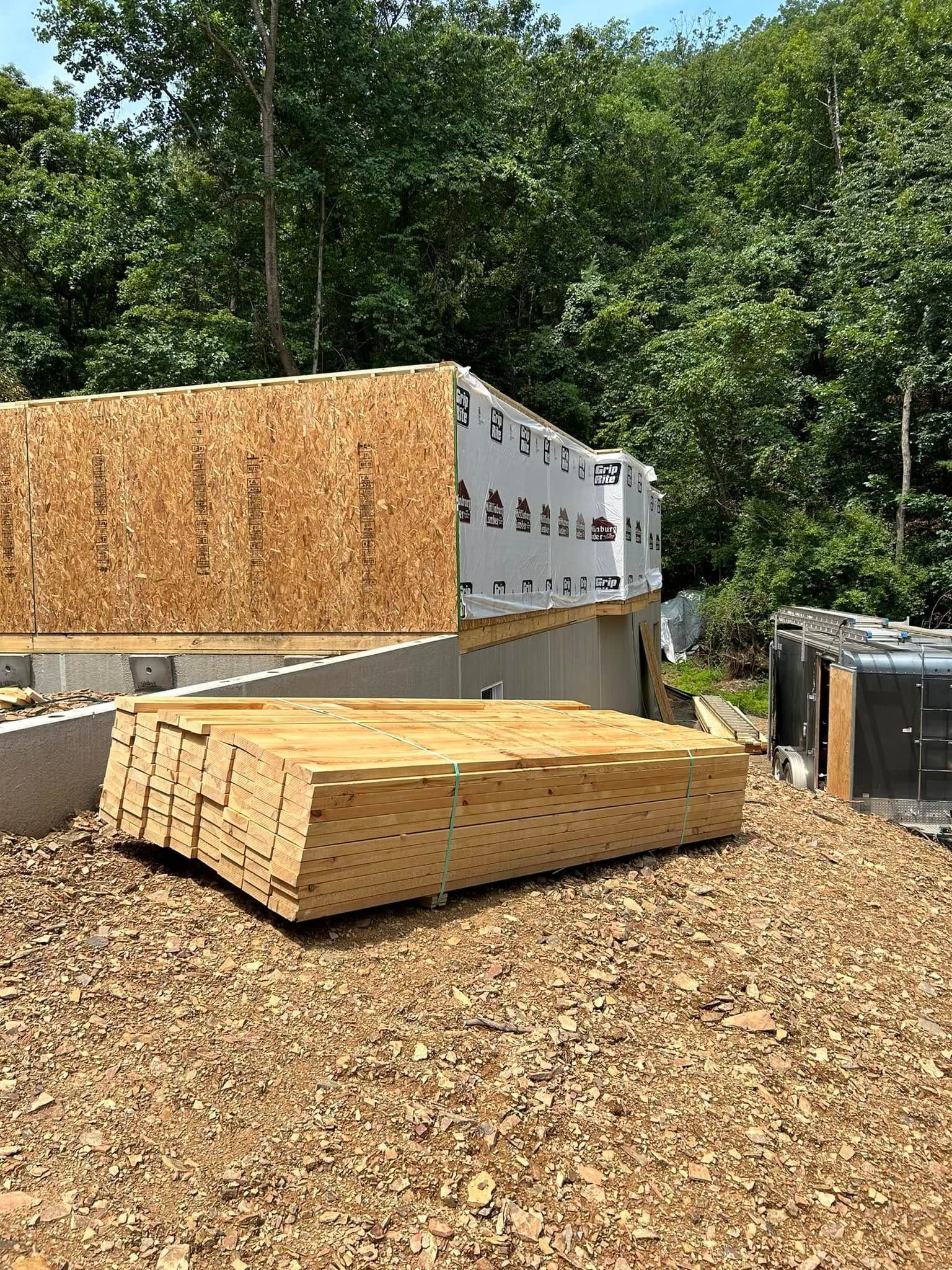 Construction site with wood-chip ground, stacked materials, and a large wrapped pallet outdoors among trees