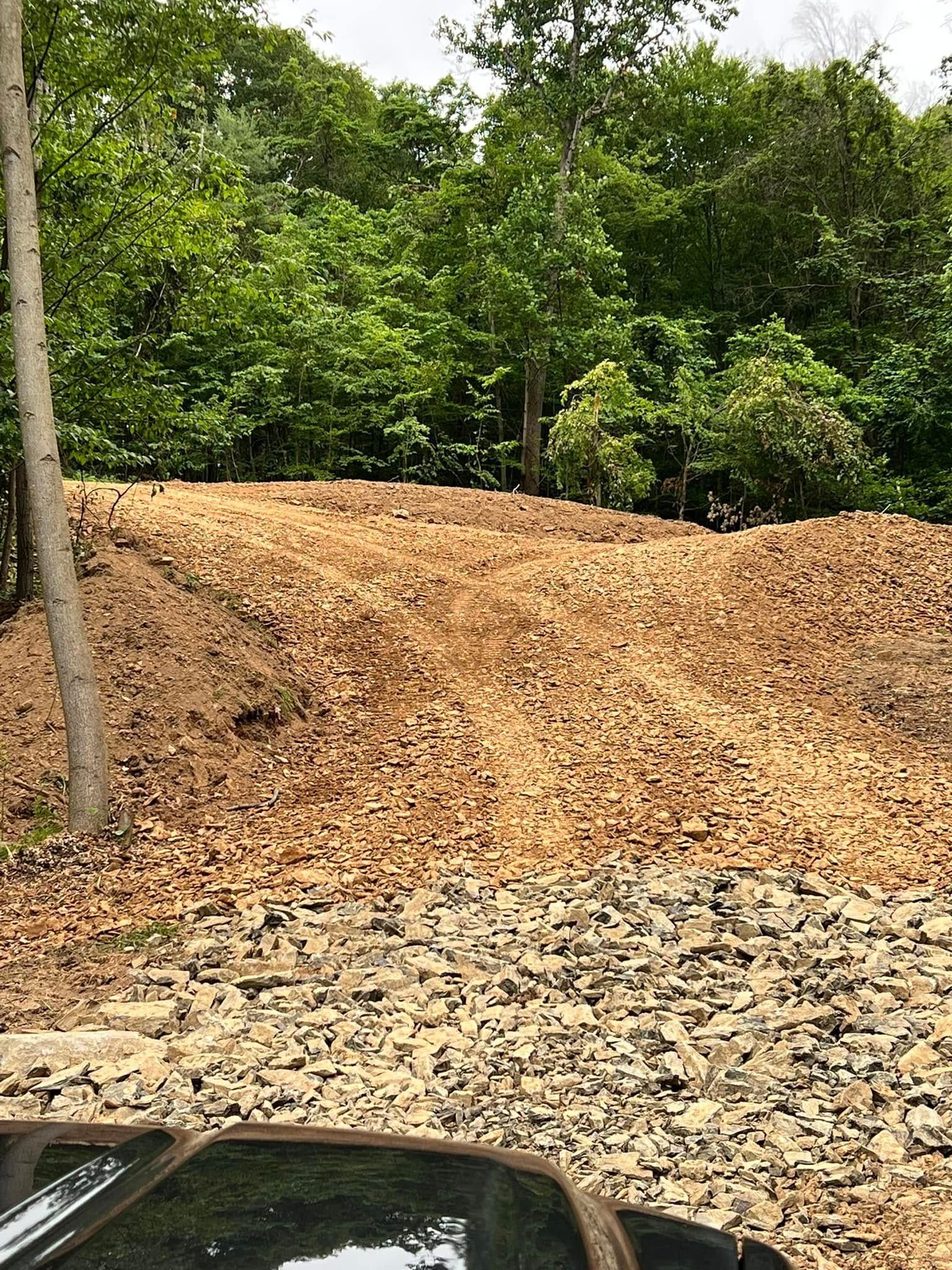 Large pile of tan gravel or dirt beside a tree-lined wooded area, with part of a dark car visible at bottom left