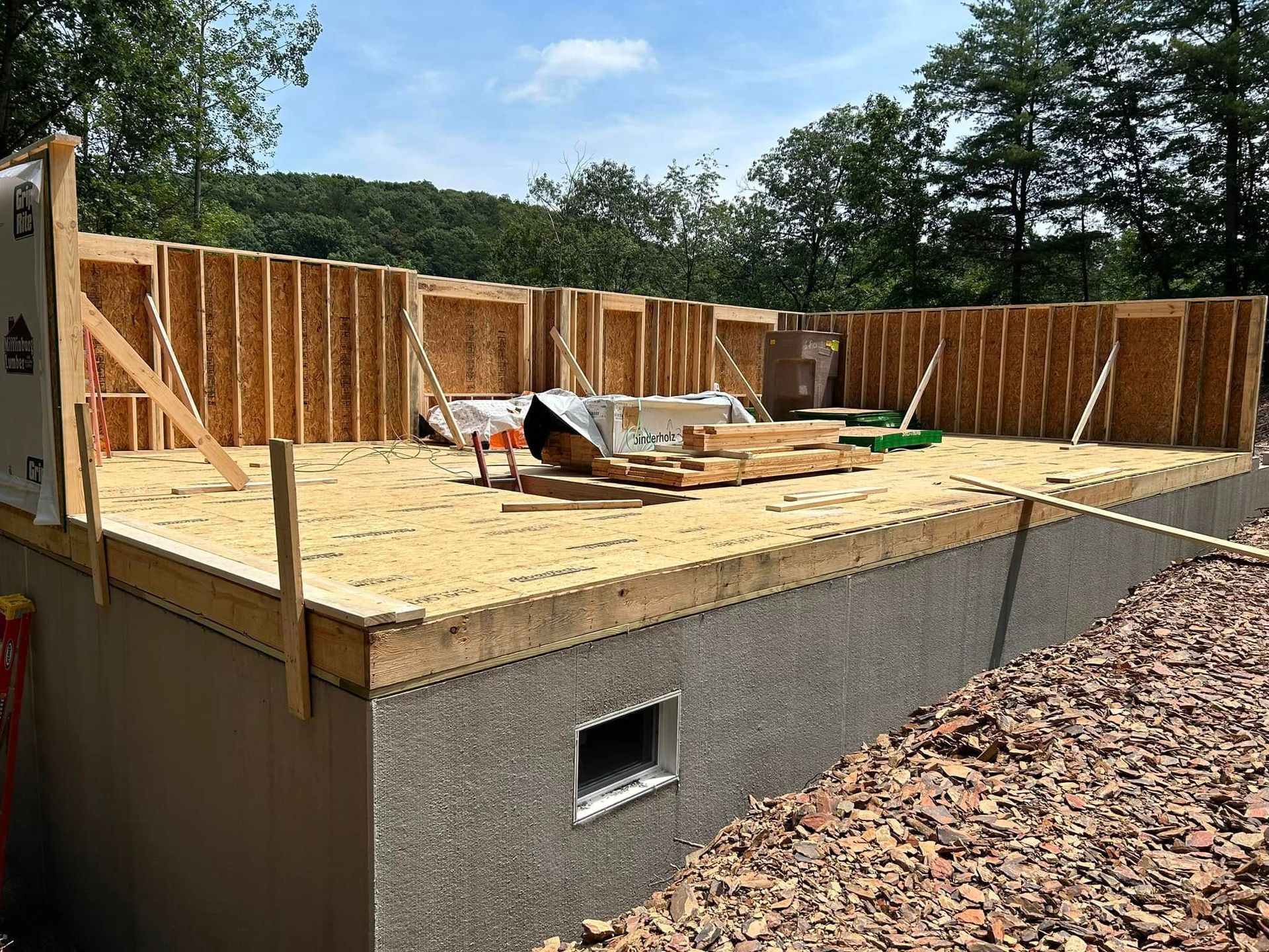 House under construction with plywood floor, framed walls, and workers on a foundation under a blue sky
