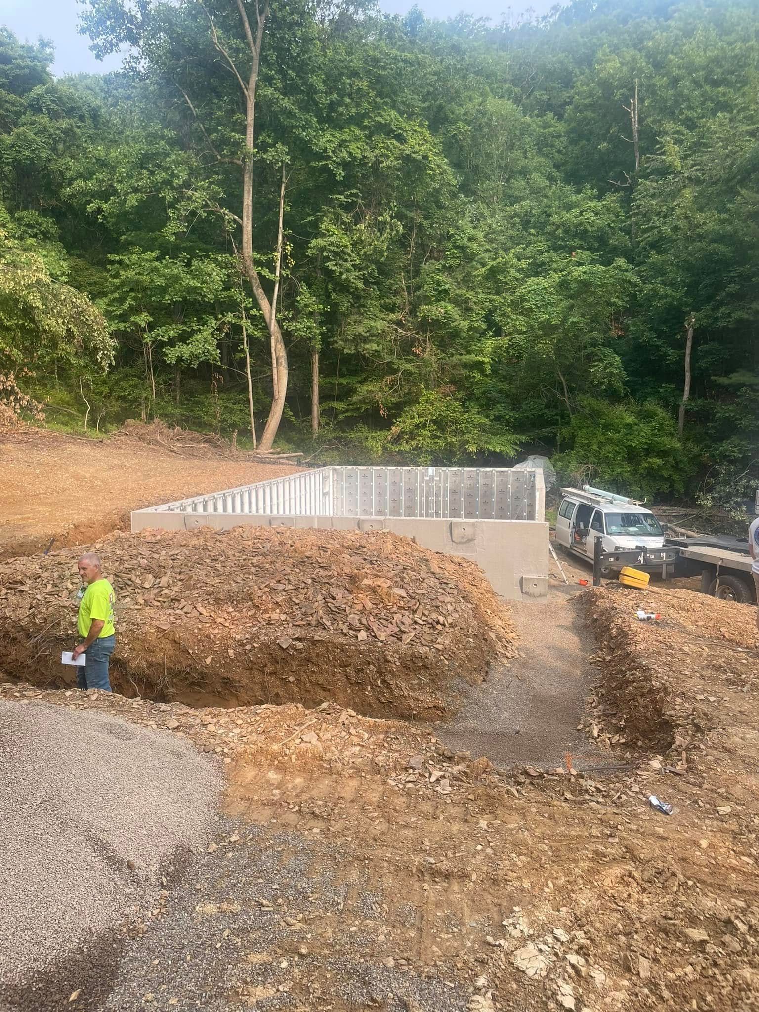 Workers on a dirt road beside a white retaining wall, with gravel piles and trees in the background.