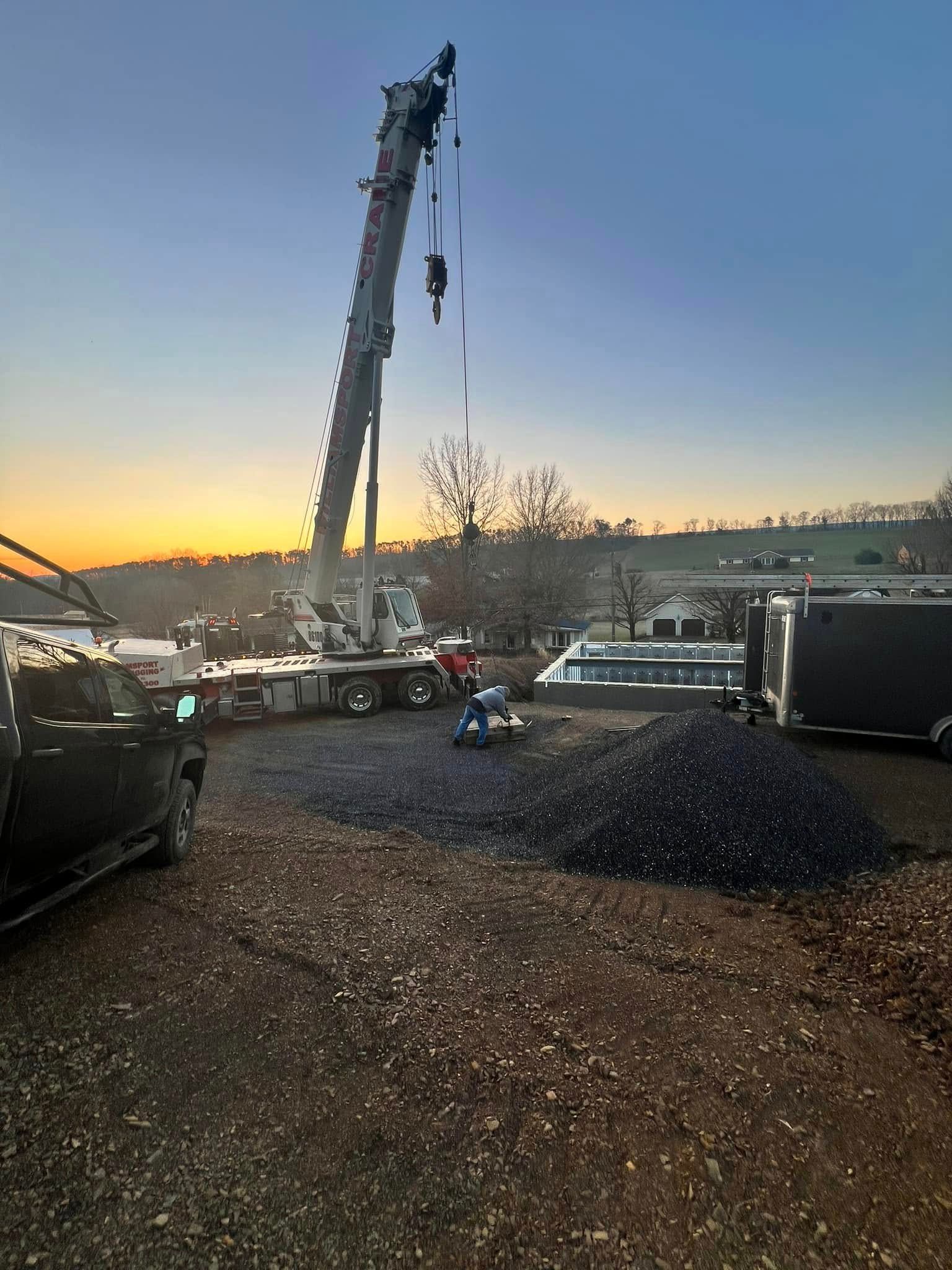 Construction crane lifting materials at dusk on a gravel lot, with vehicles and a sunset sky