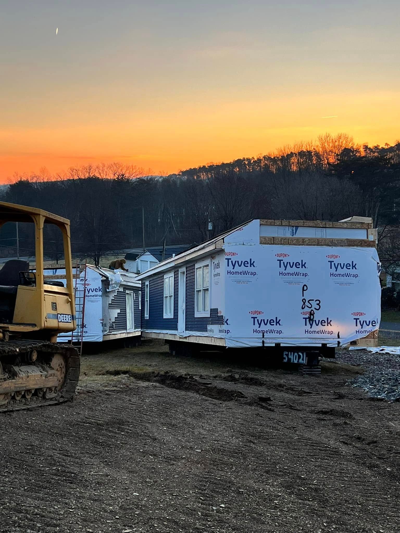Bulldozer beside a white wrapped house frame at sunset, with orange sky over trees in the background