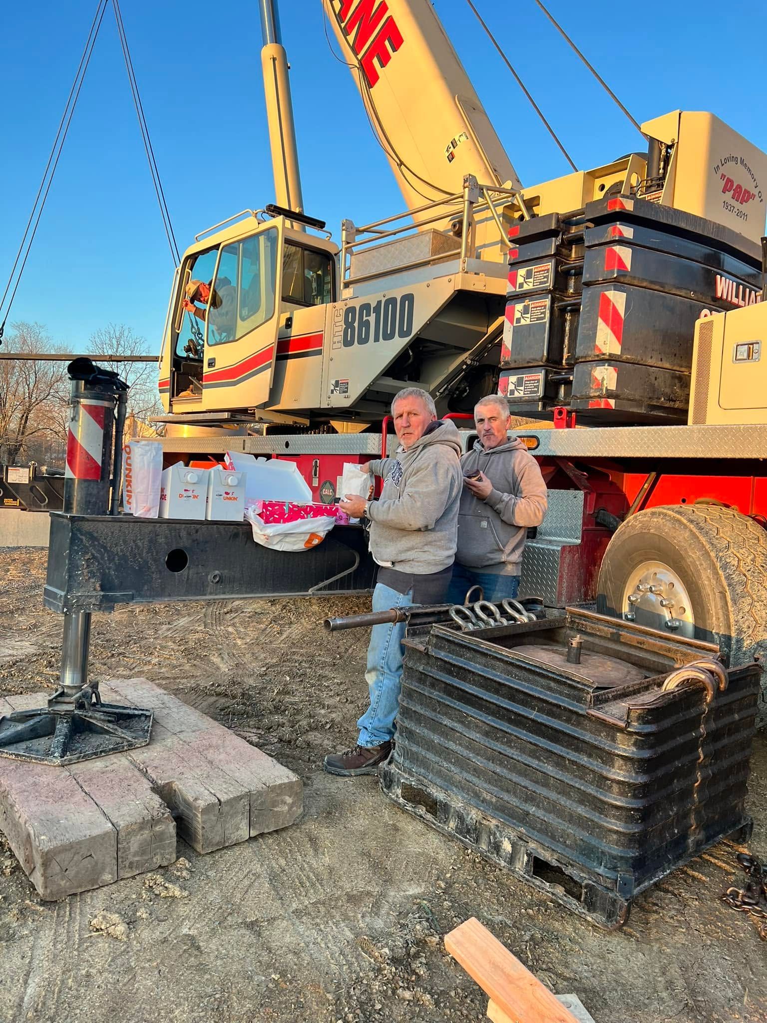 Two people posing beside a yellow construction crane and stacked concrete blocks outdoors
