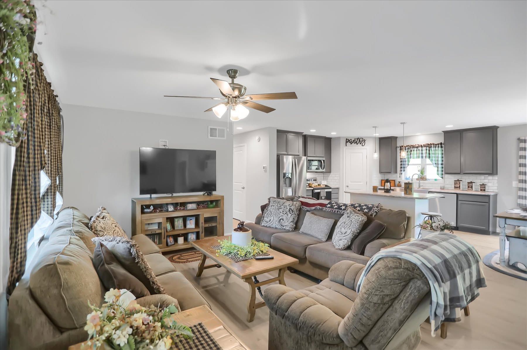 Bright living room with beige sofas, TV, ceiling fan, and open kitchen in the background