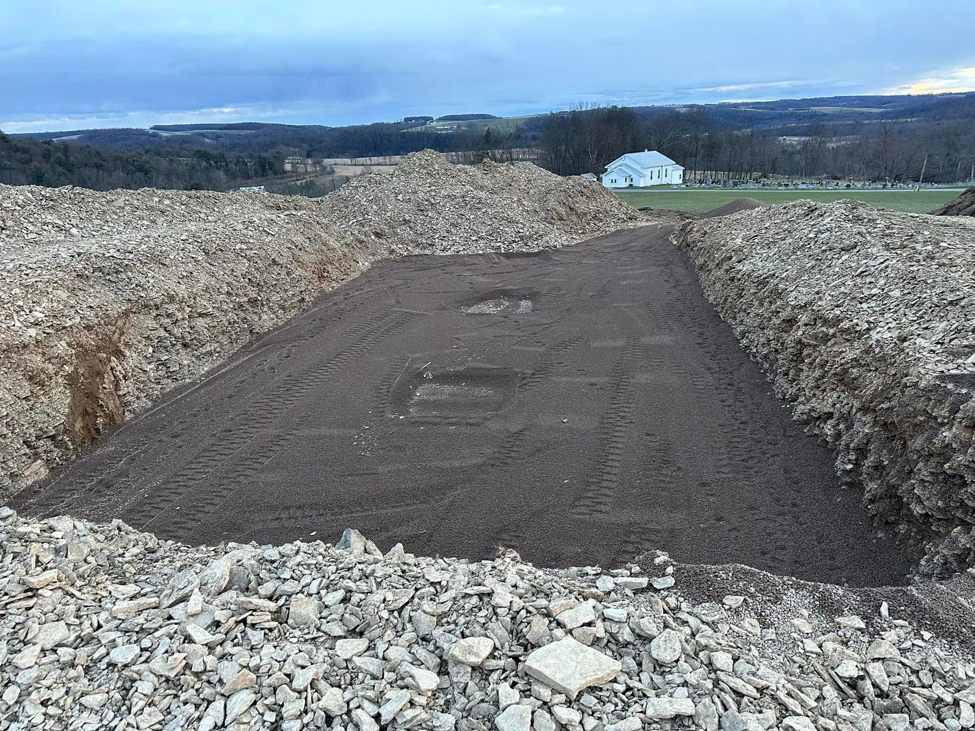 Gravel roadbed under construction through a rocky landscape, with dark soil and distant hills under a cloudy sky