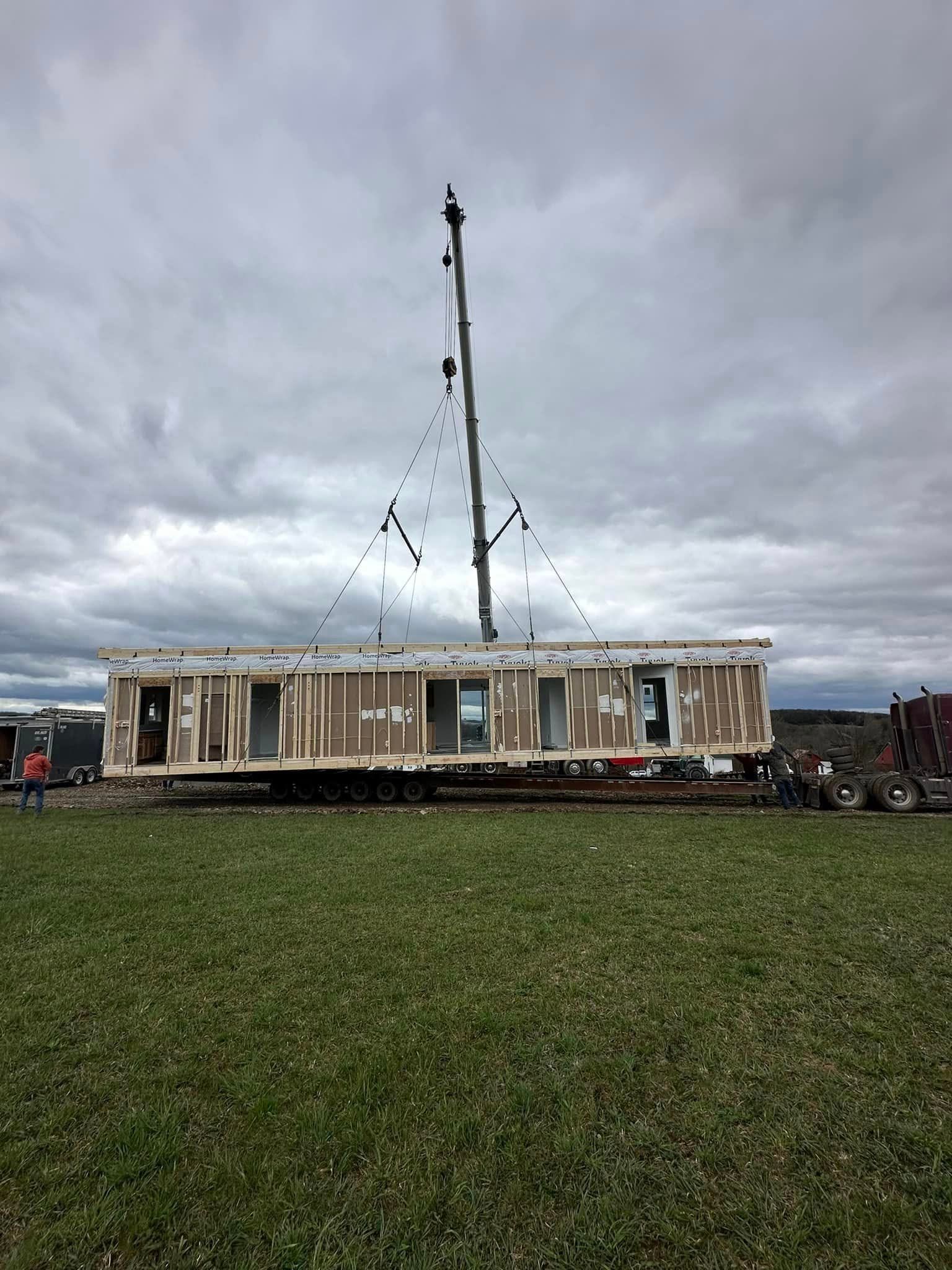 Mobile home lifted by a crane on a grassy field under a cloudy sky