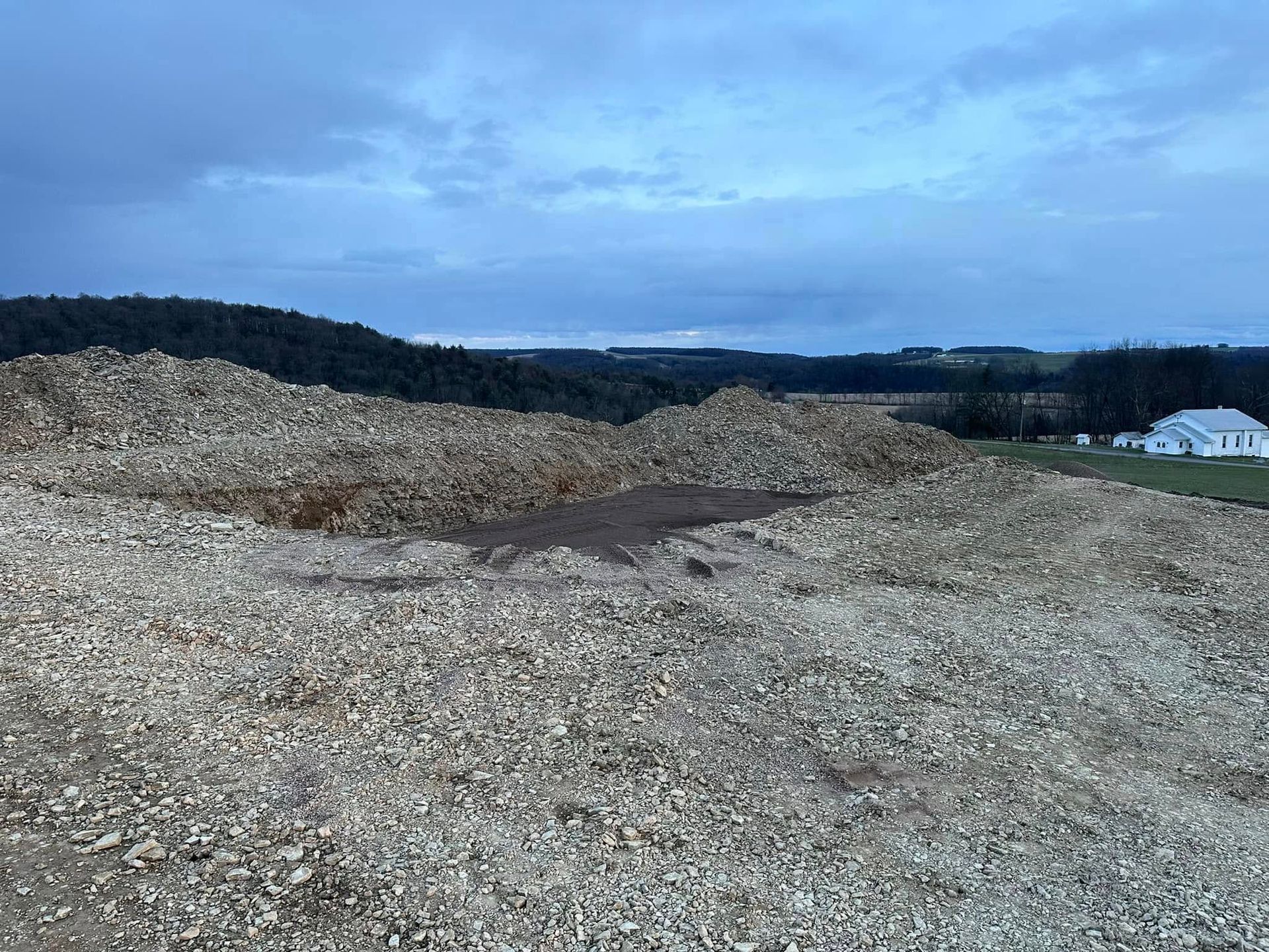 Rocky gravel mound under a cloudy blue sky, with distant hills and a white building on the right.
