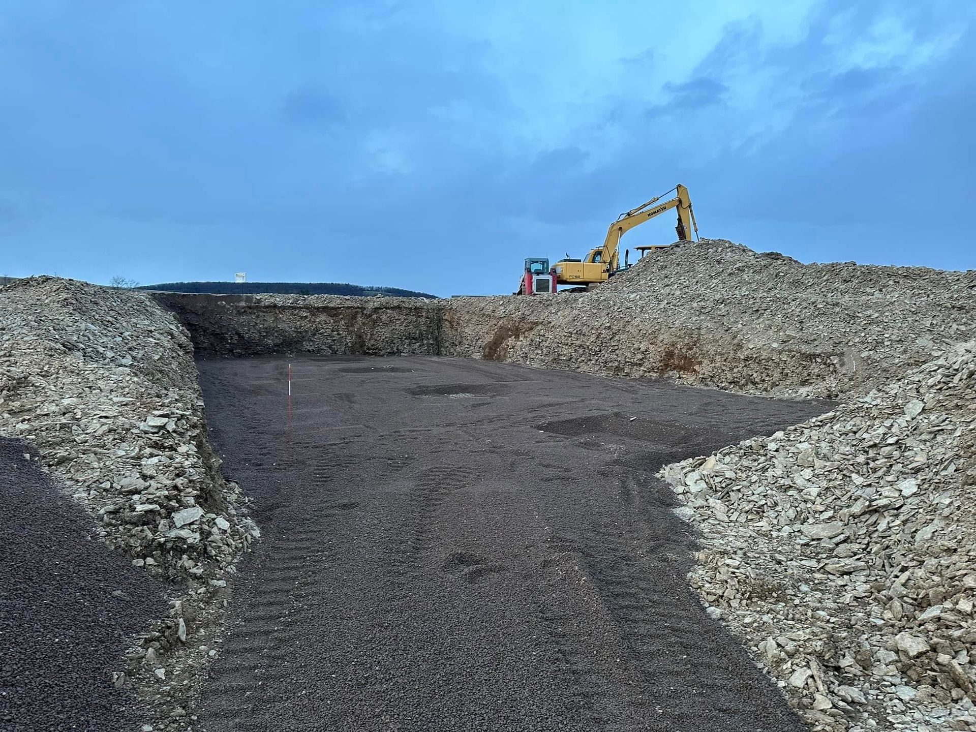 Gravel quarry road under cloudy sky, with an excavator working on a dirt mound.