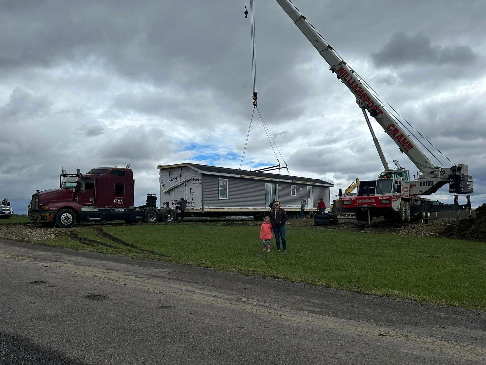 Crane lifting the roof of a mobile home beside a red truck under cloudy skies, with two people nearby