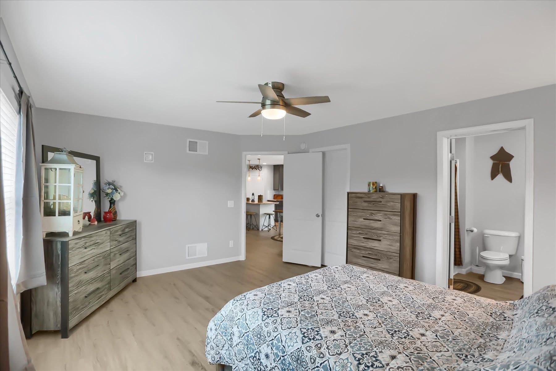 Light gray bedroom with bed, dresser, ceiling fan, and open doorway to a bathroom.