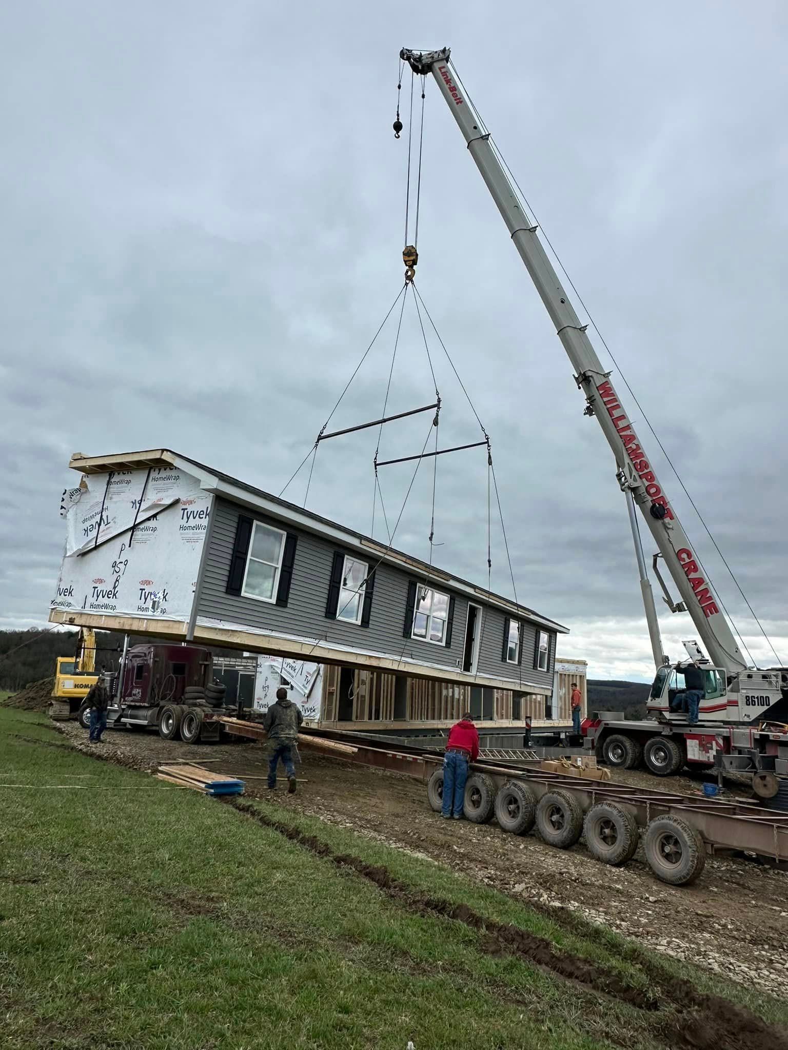 House being lifted by a crane onto a trailer, with workers nearby on a muddy lot.