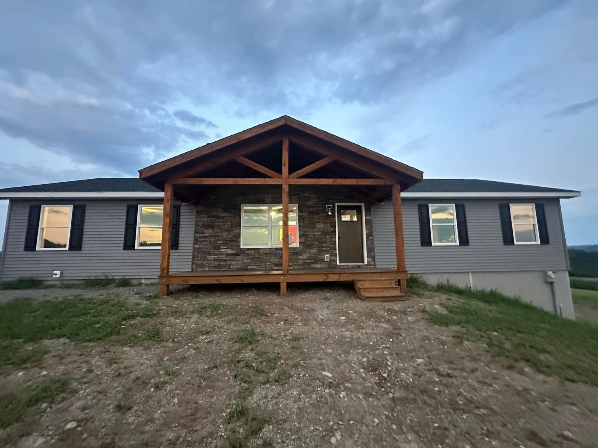 Front view of a gray house with a rustic wooden porch and gravel yard under a cloudy sky