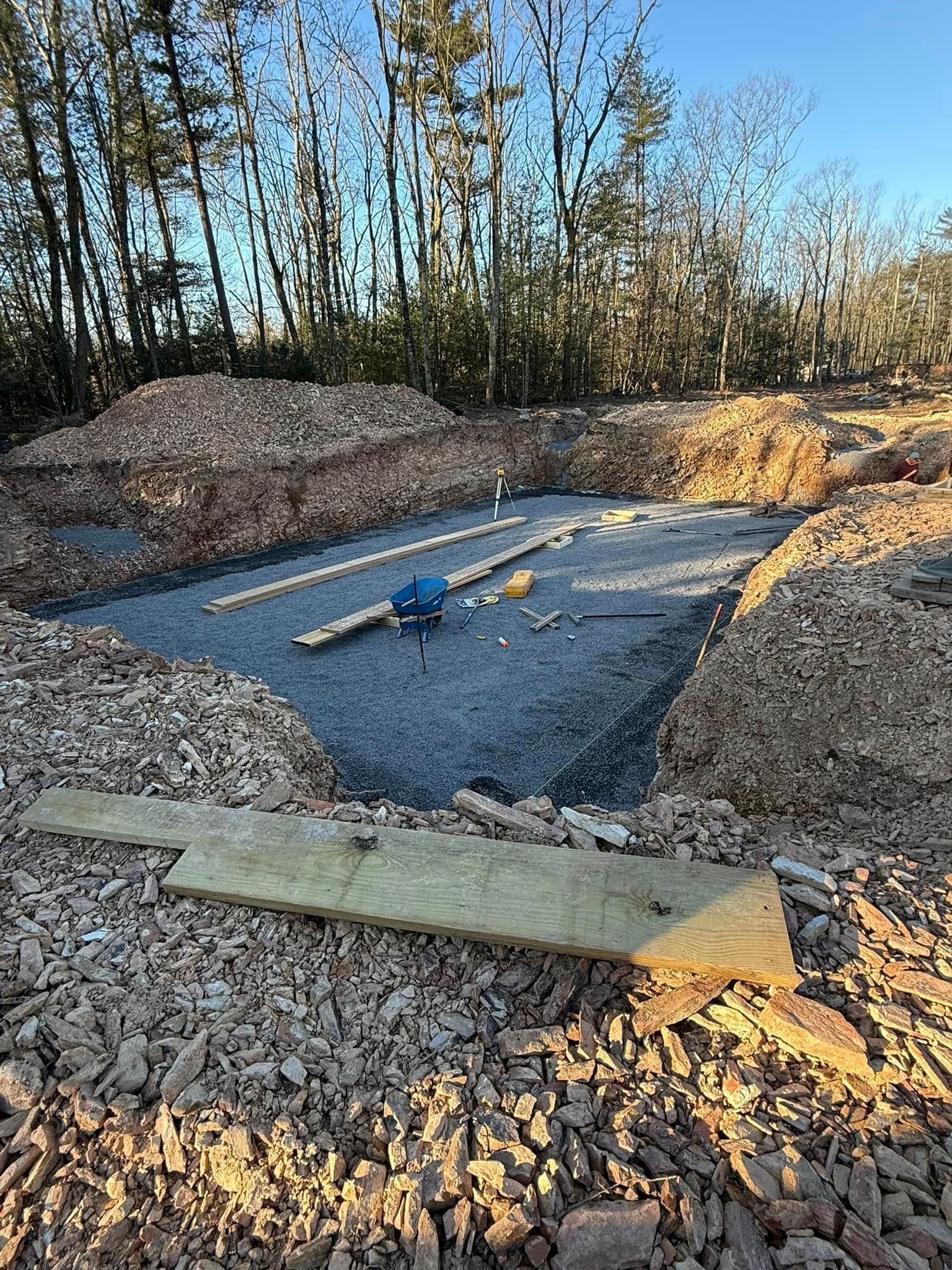 Concrete foundation slab under construction in a wooded area, with gravel, lumber, and soil piles.