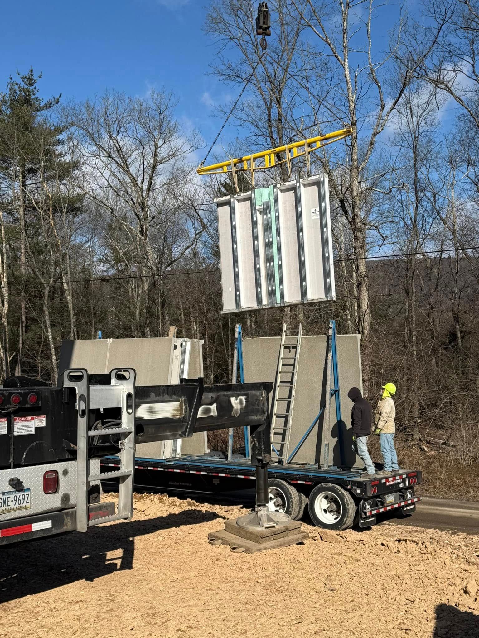 Crane lifting a large metal panel beside a truck in a wooded worksite