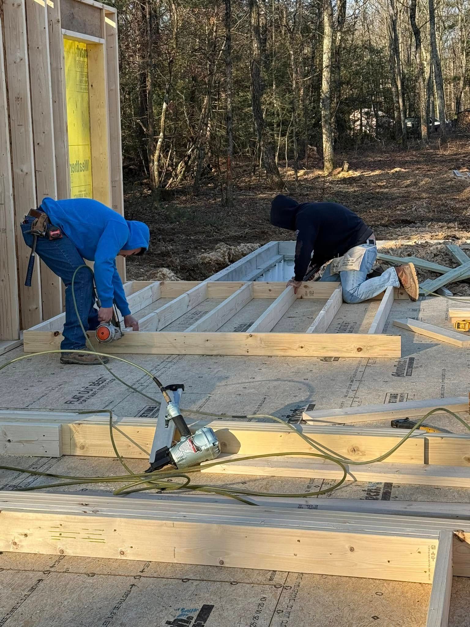 Two workers in blue and black crouch on a partially built wooden deck beside a house.