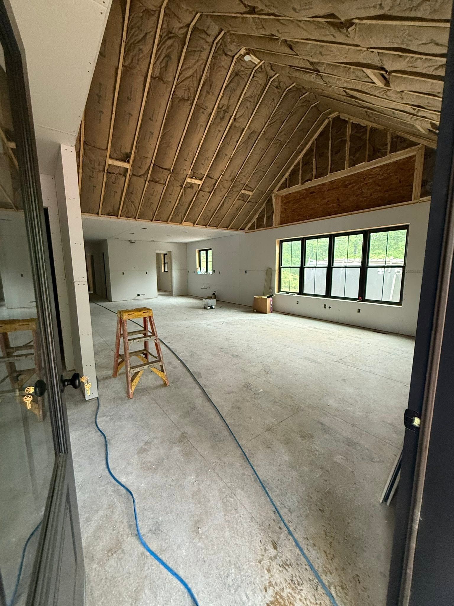 Large unfinished room with exposed ceiling beams, ladder, and windows along one wall.
