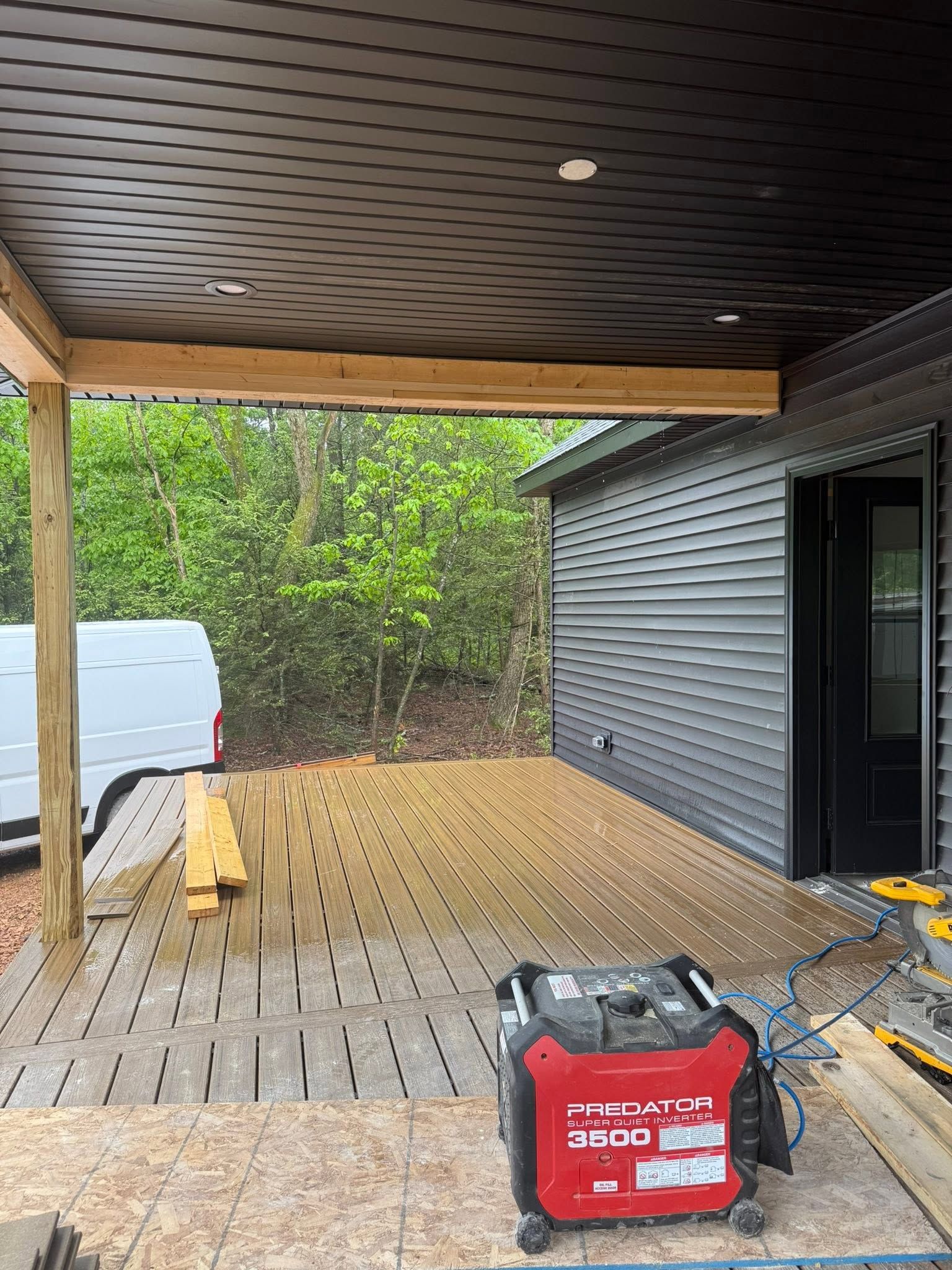 Covered wooden deck with black siding, a red tool box, and greenery beyond the railing.