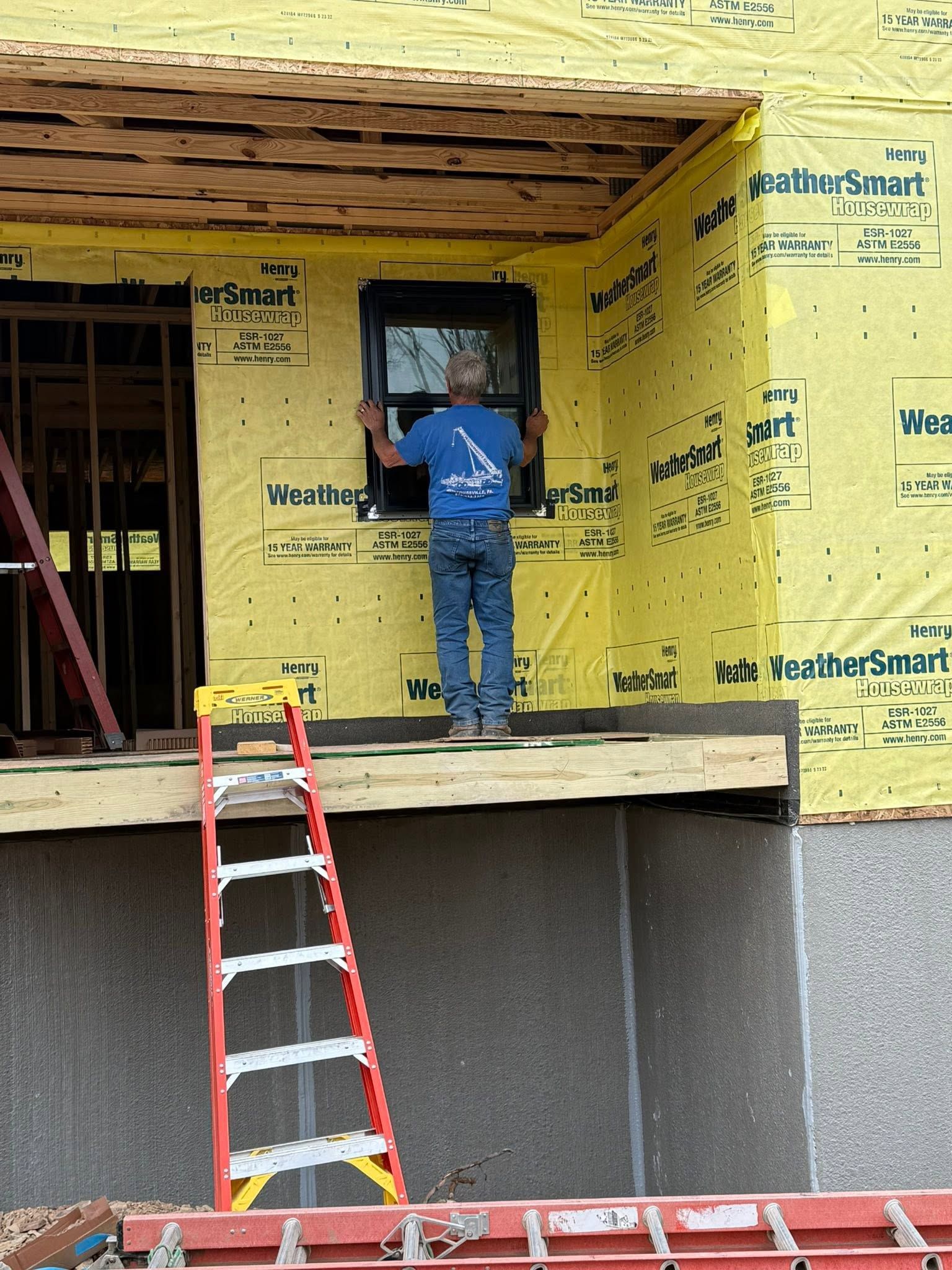 Worker in blue shirt standing on a porch, installing siding on a yellow house with a ladder below.