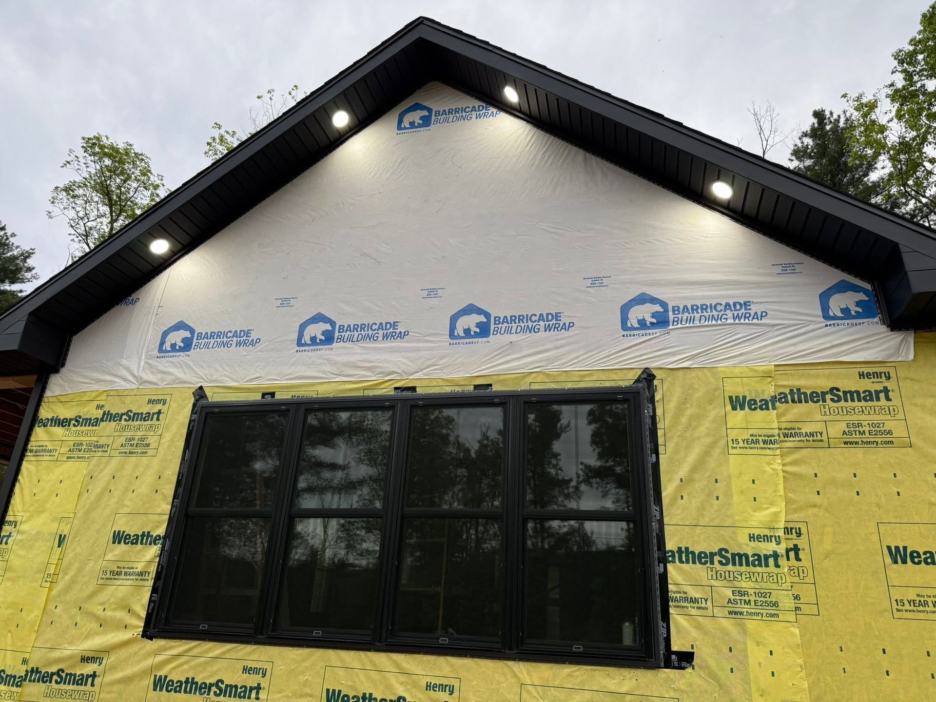 Yellow-wrapped house exterior with black gable roof and a large front window under construction