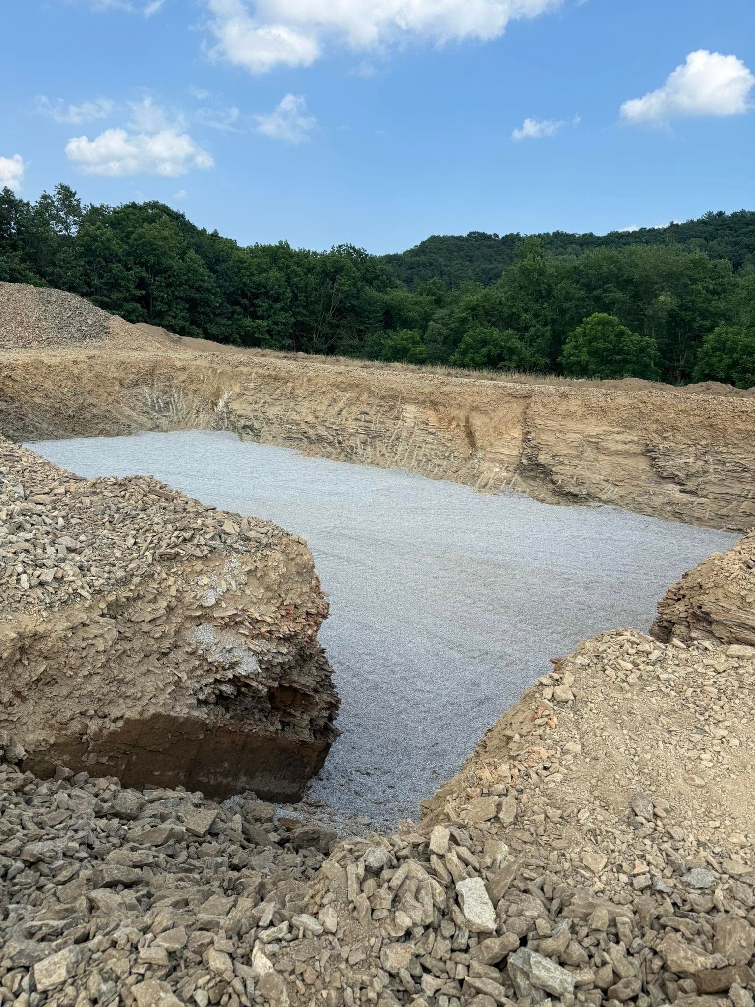Rocky quarry pit with a shallow gray water pool, surrounded by tan gravel and green trees under a blue sky