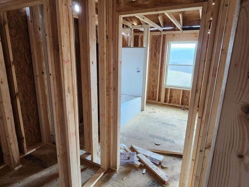 Framed wooden interior under construction with exposed studs, doorway, debris on floor, and a window at the far end