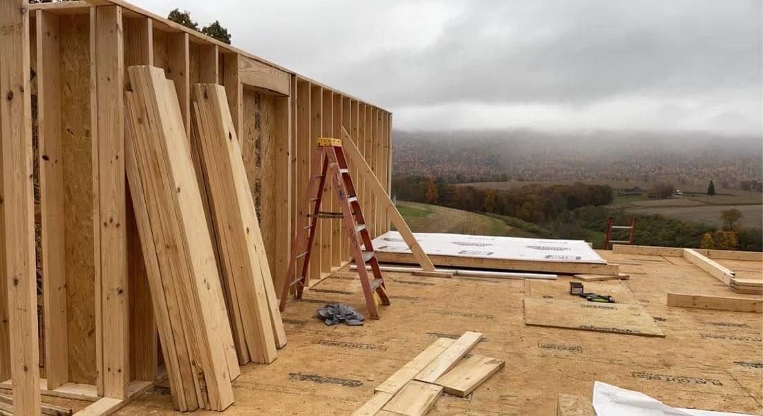 Partially framed building under construction with a red ladder on a plywood floor, under a cloudy sky