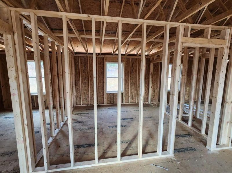 Unfinished house interior with wooden framing, exposed roof trusses, and a window at the far end.