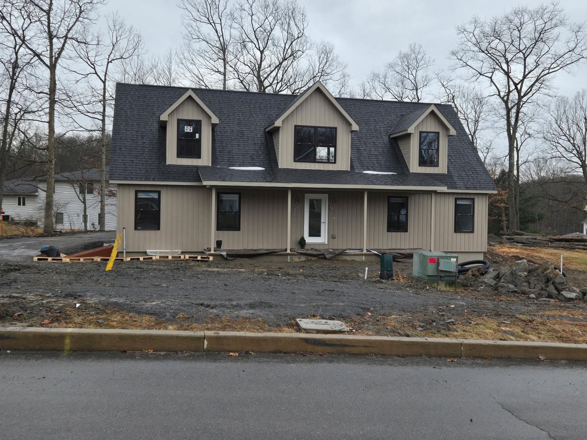 New beige house under construction with dark roof and dormer windows, front porch, and bare trees nearby