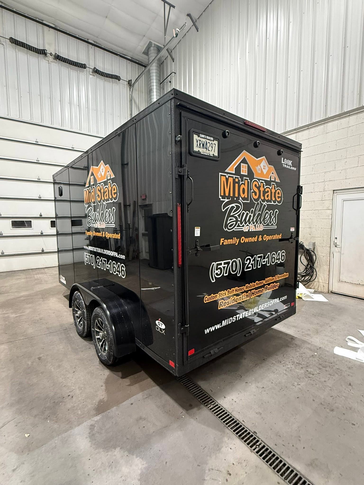 Black enclosed trailer with orange logo decals parked indoors in a white garage bay.
