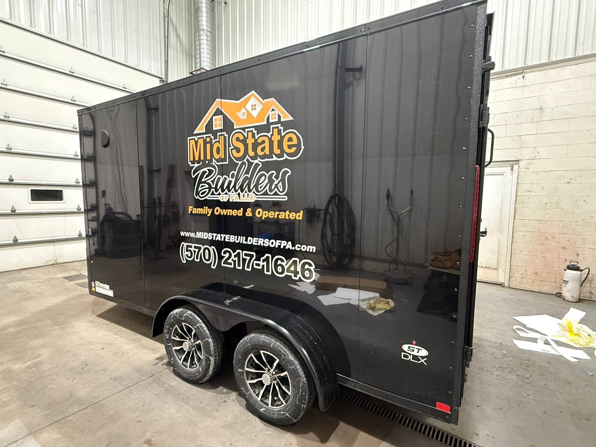 Black enclosed trailer with “Mid State Builders” logo and phone number, parked indoors on a white floor.