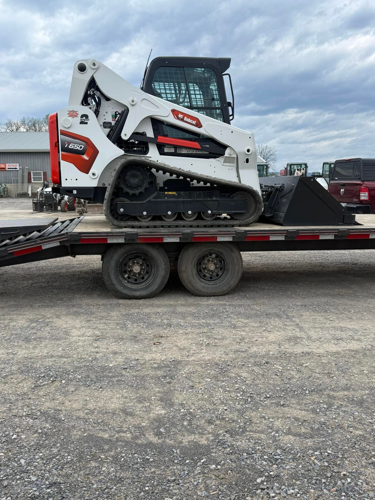 White skid steer on a red flatbed trailer in a gravel lot under cloudy skies