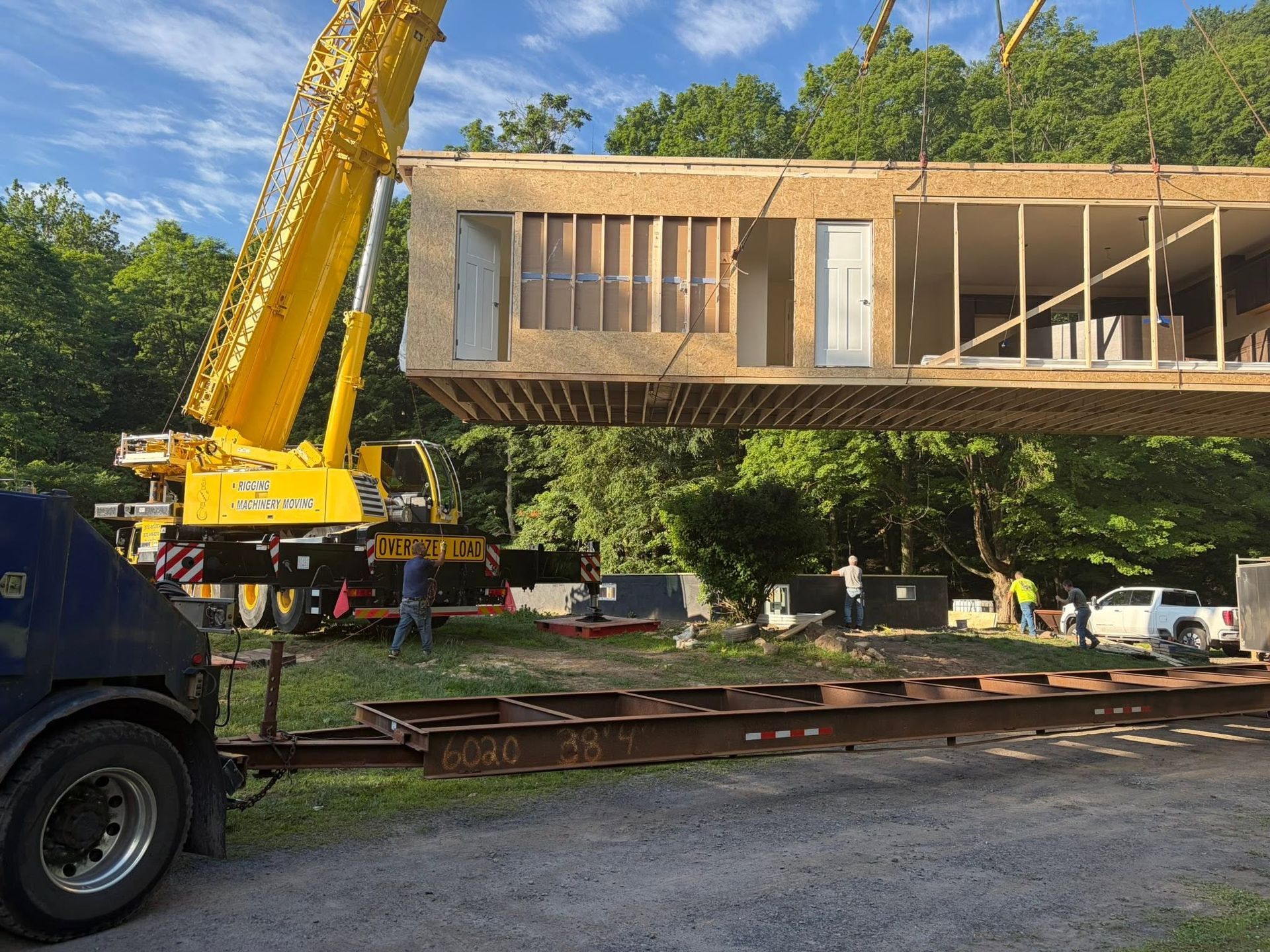 Yellow crane lifting a modern wooden house module over a flatbed truck in a wooded area