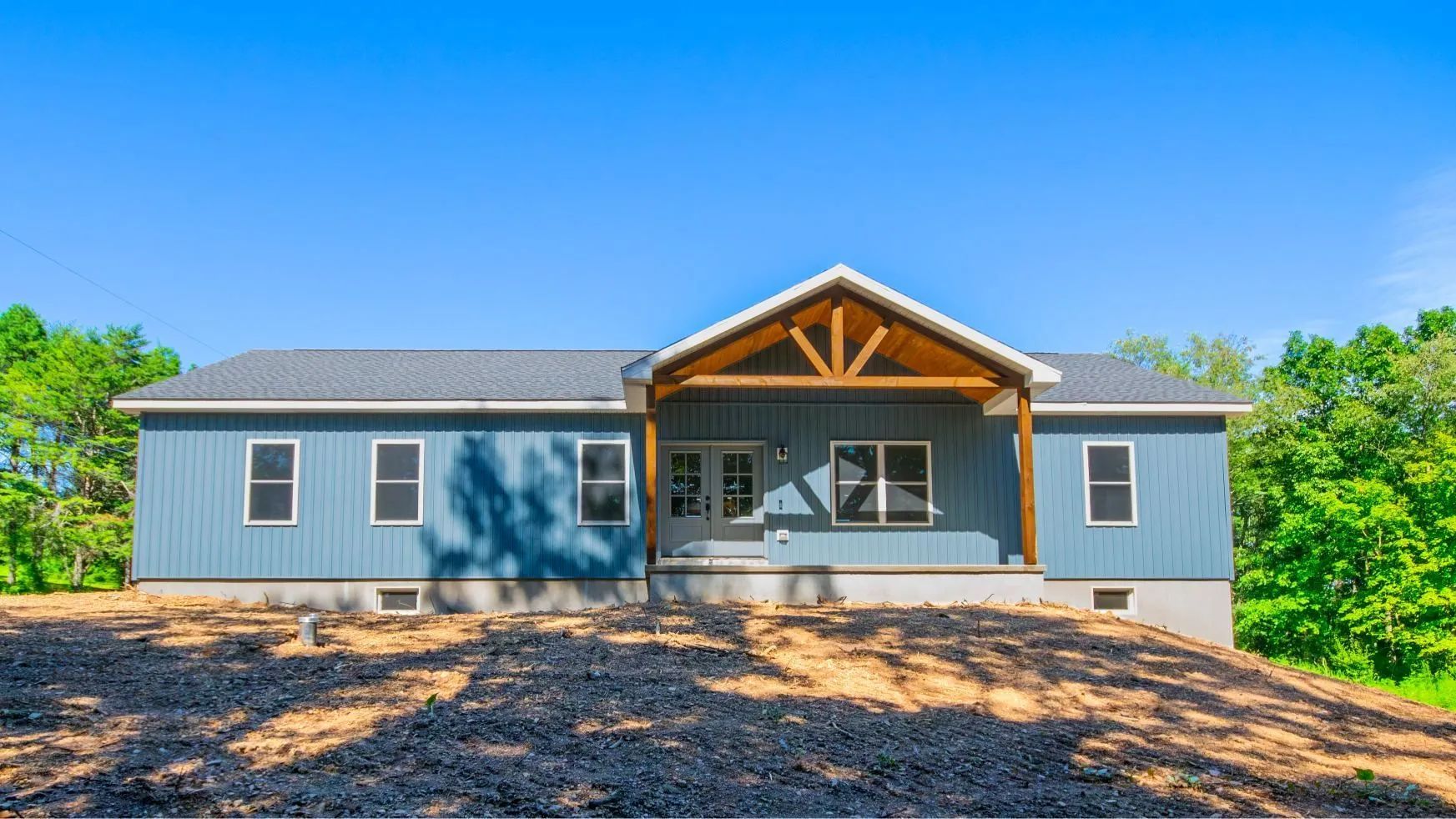 Modern gray house with a covered front porch, black roof, and wooded surroundings under a blue sky