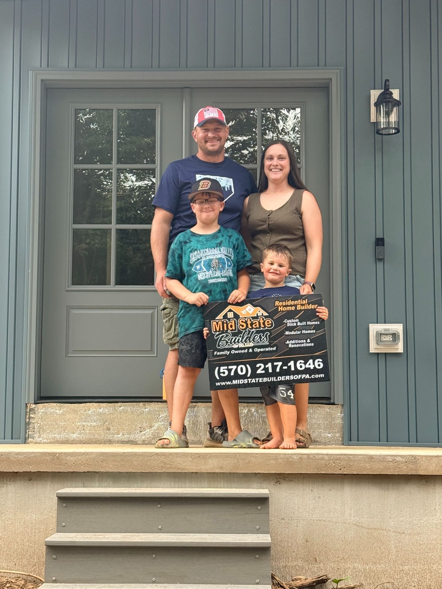 Family of four posing on a front porch, holding a real estate sign in front of a gray door.