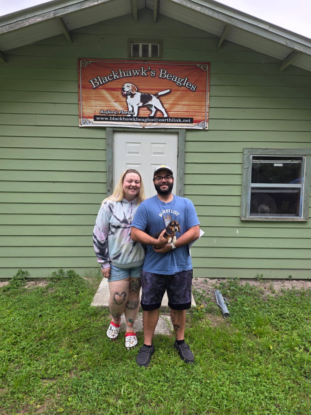 A man and a woman are standing in front of a green house holding a dog.