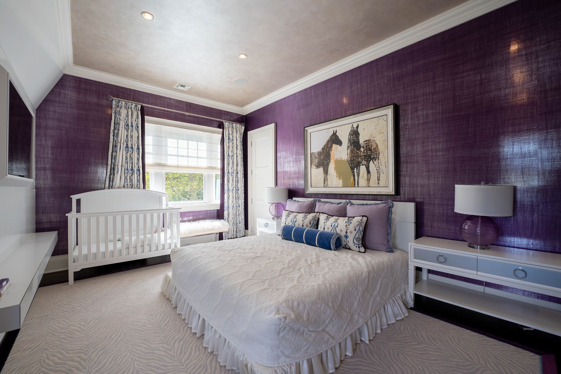 A bedroom with deep purple walls, a white bed, a baby crib, and a large horse print on the wall.