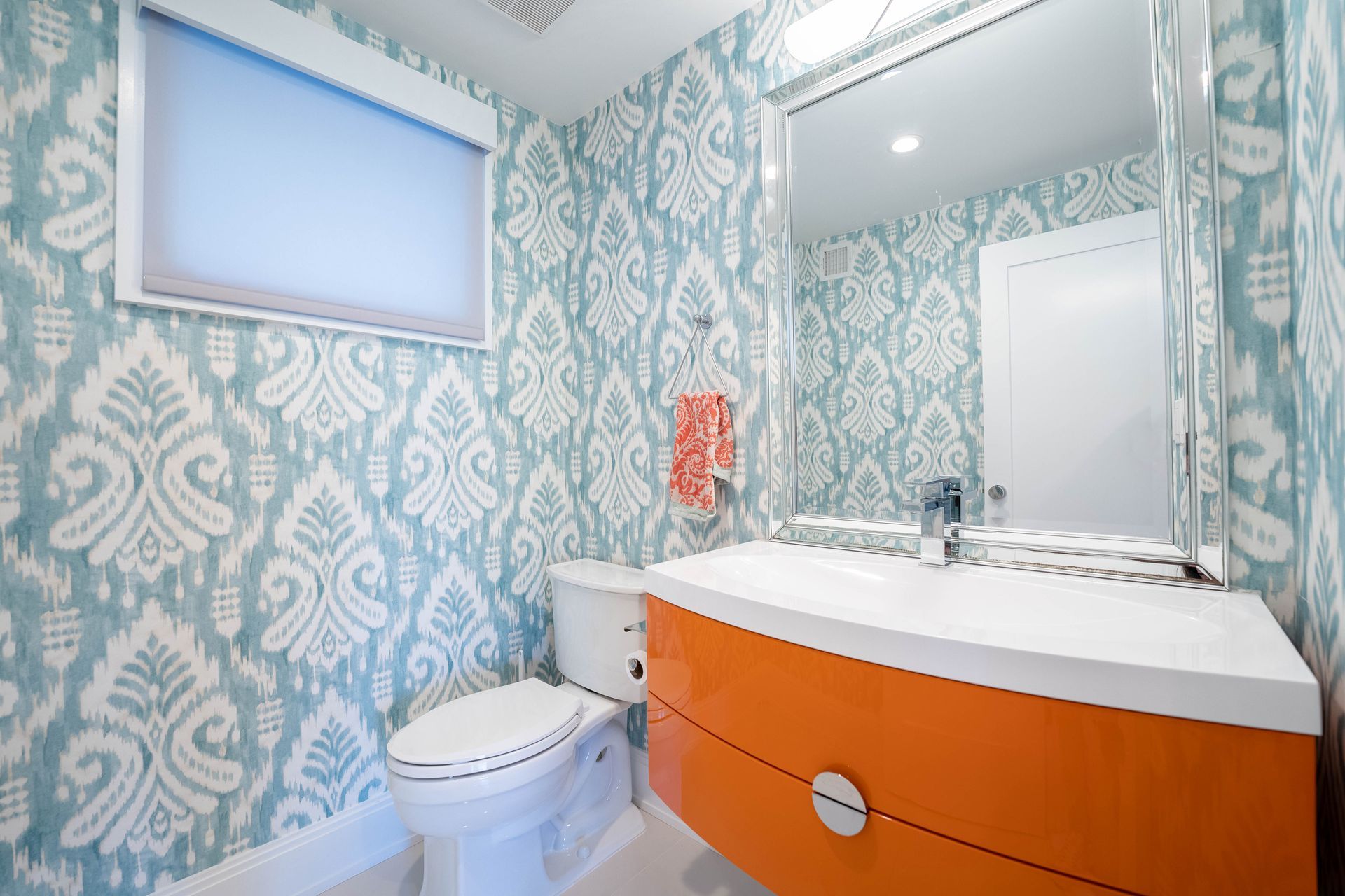 A bathroom featuring blue and white ikat wallpaper, a bright orange vanity, a white countertop, and a large mirror.