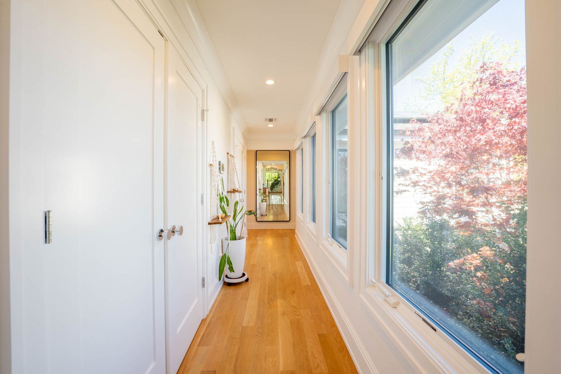 A bright hallway with wooden floors, white doors on the left, and windows on the right looking out at a red-leaved tree.