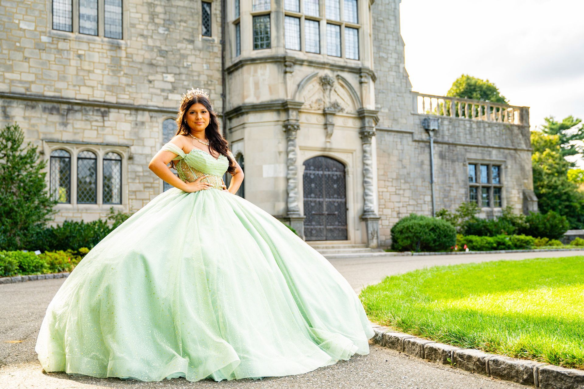 A person in a light green, full-skirted ballgown poses in front of a historic stone manor house.