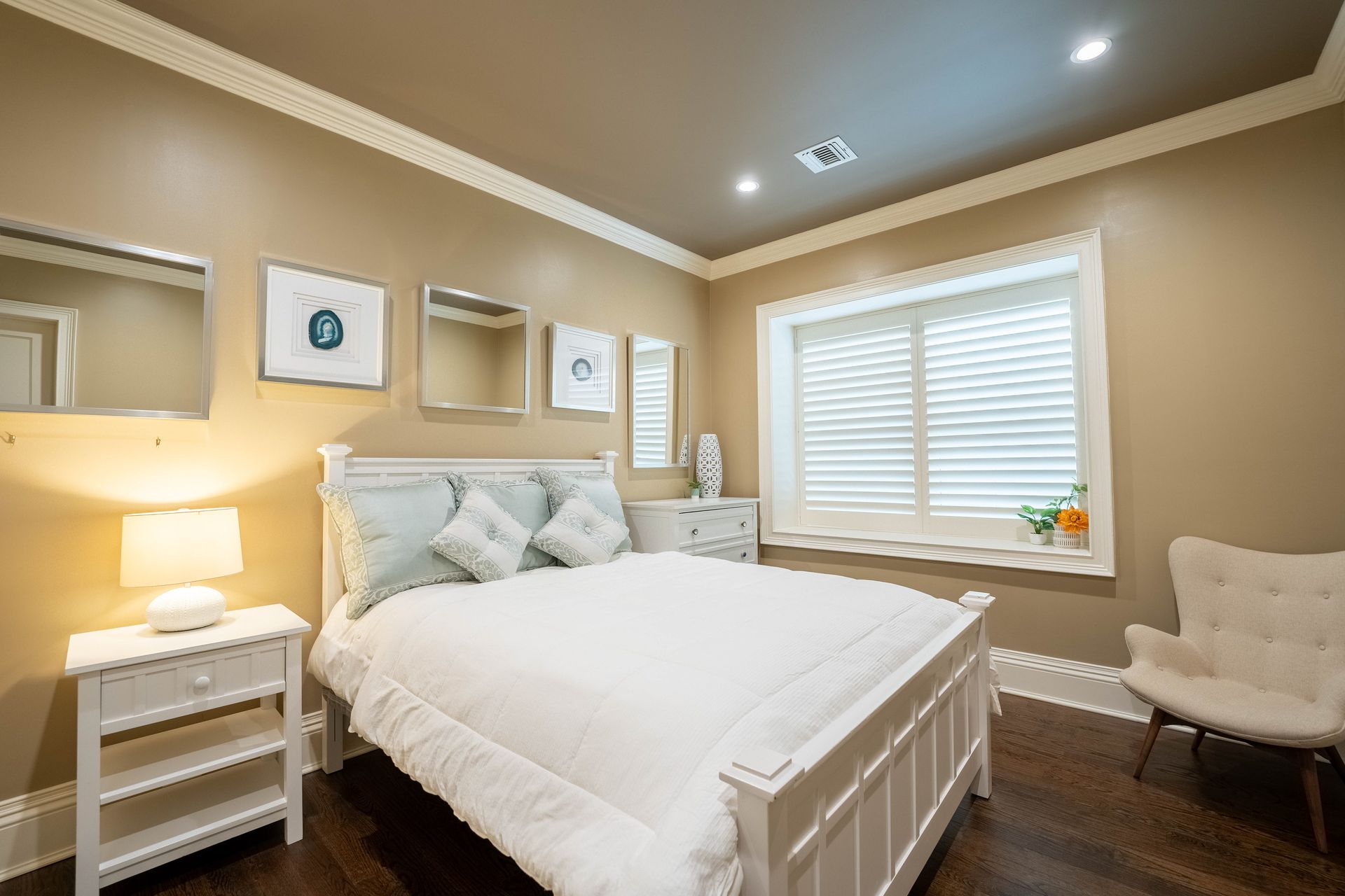 A bedroom with beige walls, a white bed, a bedside lamp, wall mirrors, framed art, and a white chair by the window.